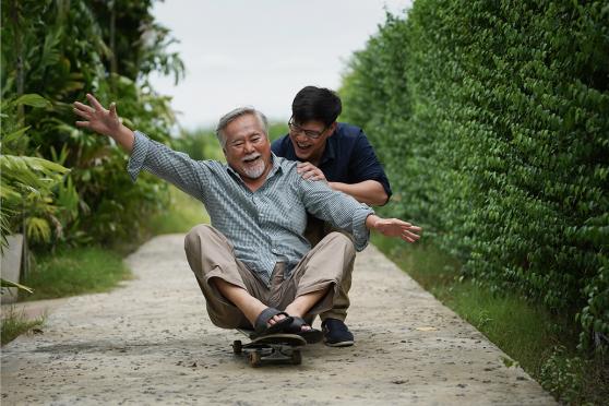 This photo shows an older man with gray hair and a beard sitting on a skateboard, laughing joyfully with his arms outstretched. A younger man with glasses and short black hair is behind him, holding onto his shoulders and smiling warmly. 